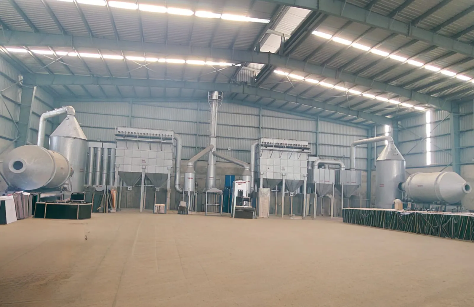 Wide workshop view of two industrial equipment trains with hopper units, cylindrical vessels, ducting and a central stack